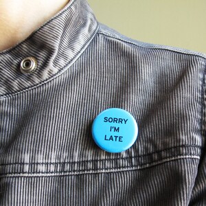 A medium blue pinback button that reads SORRY I'M LATE in plain black text. Pinned to a gray corduroy jacket, worn by a woman.
