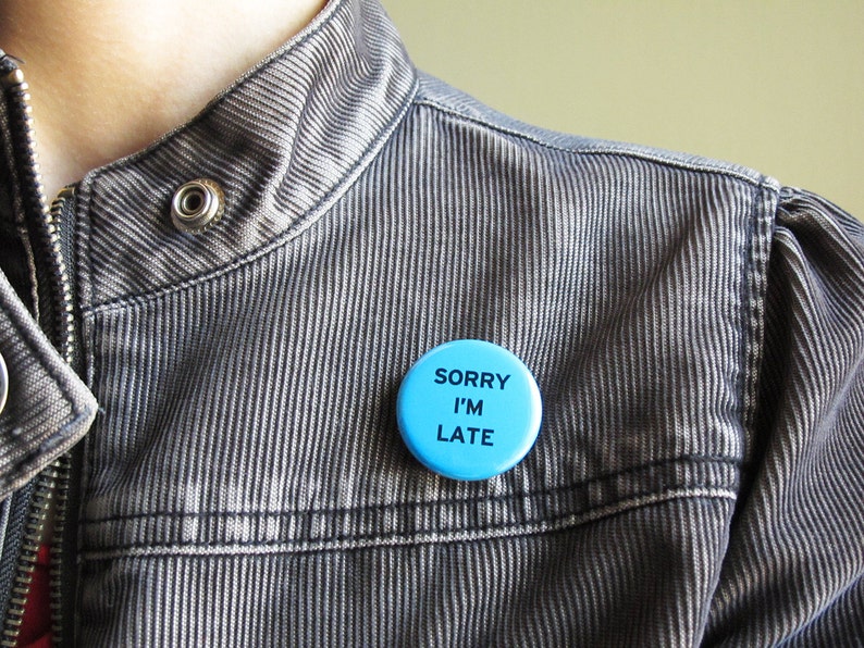 A medium blue pinback button that reads SORRY I'M LATE in plain black text. Pinned to a gray corduroy jacket, worn by a woman.