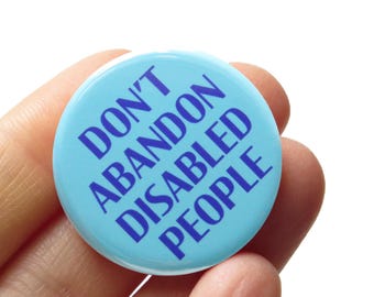 Op de afbeelding: Een ronde, lichtblauwe button met de tekst "DON'T ABANDON DISABLED PEOPLE" in donkerblauw. De button wordt in een hand gehouden tegen een witte achtergrond. De button is een draagbaar accessoire.