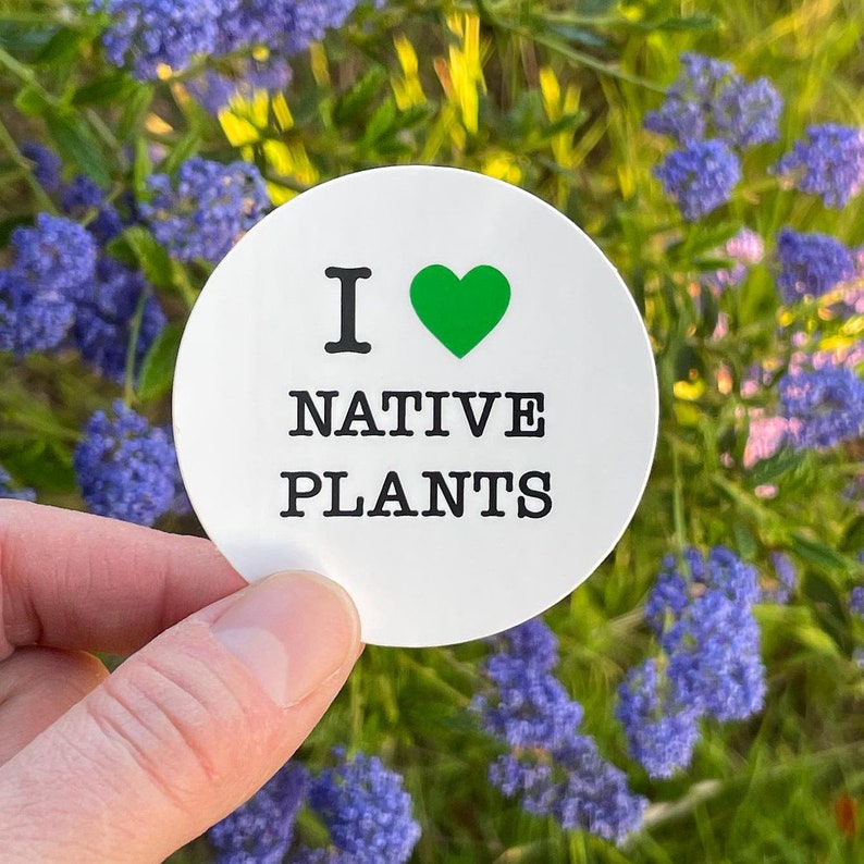 A round white sticker that reads I LOVE NATIVE PLANTS, love being indicated by a green heart. Sticker is held in the corner in front of greenery and flowers, a wild native plants garden.