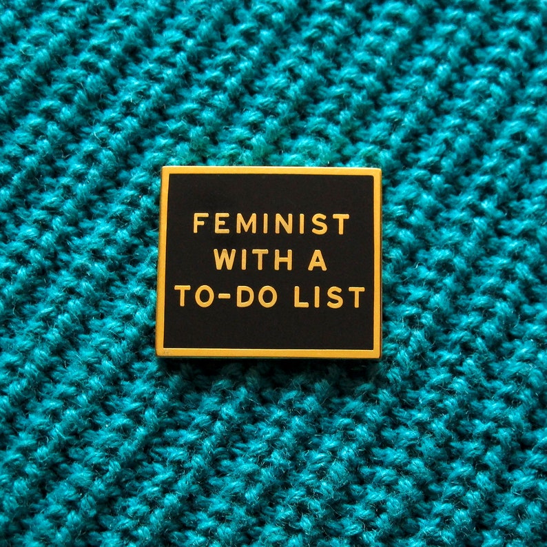 A square rectangle enamel pin with gold outline & text on a black enamel background, reading FEMINIST WITH A TO-DO LIST. Pin is on a diagonal weave teal cotton sweater.