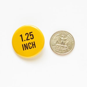 May include: A circular yellow button with the black text 1.25 INCH printed on the front, positioned next to a United States quarter coin for scale, demonstrating the button has a diameter of 1.25 inches as indicated by the printed text.