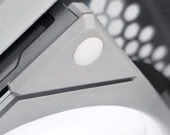 May include: A computer cooling fan featuring a light grey frame with a circular white illuminated centre. The fan assembly includes a corner mounting point with a small circular accent and a visible honeycomb-patterned ventilation grille nearby.