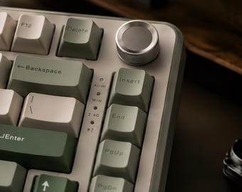 May include: A mechanical keyboard featuring a colour scheme of off-white and sage green keycaps. The layout includes a circular silver volume knob in the top right corner, with indicator lights for connection modes labelled as AU/IN, WIN, 2.4G, and BT.