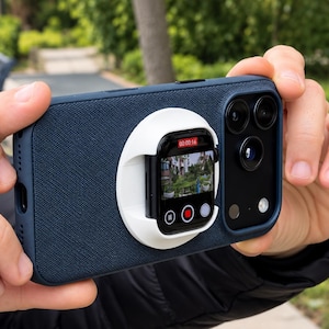 以下が含まれることがあります： ダークブルーのテクスチャ加工されたスマートフォンケースで、背面に円形の白いマウントが取り付けられています。マウントには黒いスマートウォッチが固定されており、画面には赤い録画インジケーターと再生アイコンが表示されたライブカメラ映像が映し出されています。