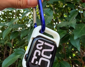 May include: A white protective case with a blue carabiner clip holding a black smartwatch. The watch face displays the time in white numerals against a black background. The case is held against a backdrop of green foliage.