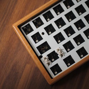 May include: A mechanical keyboard case made of light-toned wood featuring a white mounting plate with a grid of square cutouts. Two white stabilizer mechanisms are installed in the lower section of the exposed internal keyboard layout.