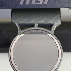 Op de afbeelding: Een zwarte 3D-geprinte plastic monitorbevestiging met een cirkelvormige magnetische voorplaat. De houder is bevestigd aan de onderkant van een donker monitorframe met het witte MSI-logo op de onderste rand van de schermrand.