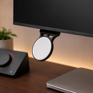 Op de afbeelding: Een zwarte ronde magnetische houder bevestigd aan de onderrand van een computermonitor. De houder heeft een plat, lichtgrijs rond oppervlak en een zwart frame dat naar boven uitsteekt om aan de rand van de monitor te worden bevestigd.