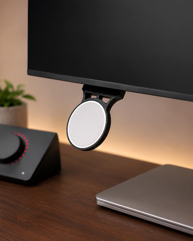 Op de afbeelding: Een zwarte ronde magnetische houder bevestigd aan de onderrand van een computermonitor. De houder heeft een plat, lichtgrijs rond oppervlak en een zwart frame dat naar boven uitsteekt om aan de rand van de monitor te worden bevestigd.