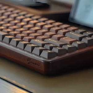 May include: A mechanical keyboard featuring a dark brown wooden frame and dark brown keycaps with gold-colored legends. The front edge of the wooden case displays a cursive engraved signature. The keys are arranged in a standard layout with a slight angle.