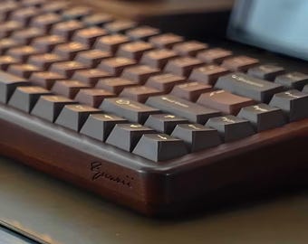 May include: A mechanical keyboard featuring a dark brown wooden frame and dark brown keycaps with gold-coloured legends. The front edge of the wooden case displays a cursive engraved signature. The keys are arranged in a standard layout with a slight angle.