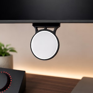 May include: A circular white magnetic mount with a black frame attached to the bottom edge of a black computer monitor. The device features a smooth, flat surface and a sturdy bracket that secures it directly to the display frame for accessory use.
