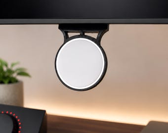 May include: A circular white magnetic mount with a black frame attached to the bottom edge of a black computer monitor. The device features a smooth, flat surface and a sturdy bracket that secures it directly to the display frame for accessory use.