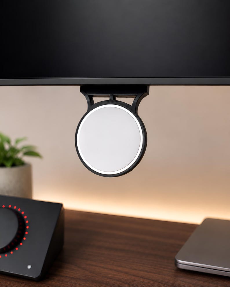 Op de afbeelding: Een cirkelvormige witte magnetische houder met een zwart frame bevestigd aan de onderrand van een zwarte computermonitor. Het apparaat heeft een glad, plat oppervlak en een stevige beugel die direct aan het schermframe wordt bevestigd.