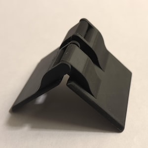 May include: A black plastic hinge with two interlocking parts. The hinge is designed to fold at a 90-degree angle.
