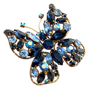May include: A butterfly-shaped brooch with a gold-tone frame. The brooch is adorned with various shades of blue and clear crystals. The crystals are in different shapes and sizes, creating a sparkling effect.