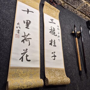 May include: Two traditional Asian scroll paintings with calligraphy in black ink. The scrolls have gold patterned borders and are hanging on a dark gray surface. Two calligraphy brushes are also visible.
