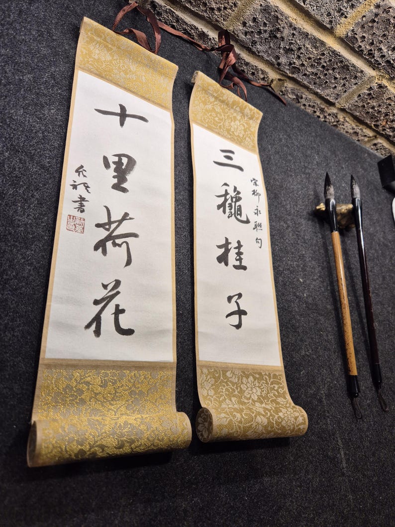 May include: Two traditional Asian scroll paintings with calligraphy in black ink. The scrolls have gold patterned borders and are hanging on a dark gray surface. Two calligraphy brushes are also visible.