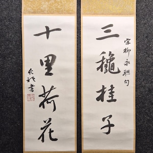 May include: Two traditional Chinese calligraphy scrolls. The scrolls feature black ink characters on white paper, with gold patterned borders and brown ribbons for hanging. The text is in vertical columns, with a red seal on one scroll.