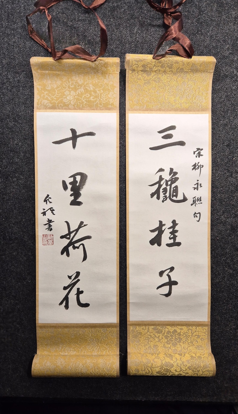May include: Two traditional Chinese calligraphy scrolls. The scrolls feature black ink characters on white paper, with gold patterned borders and brown ribbons for hanging. The text is in vertical columns, with a red seal on one scroll.