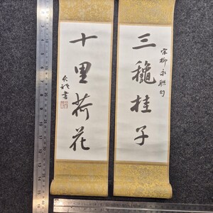 May include: Two traditional Asian scroll paintings. Each scroll features calligraphy on white paper, framed by gold patterned borders. The scrolls are suspended by brown ribbons. A ruler is placed next to the scrolls.