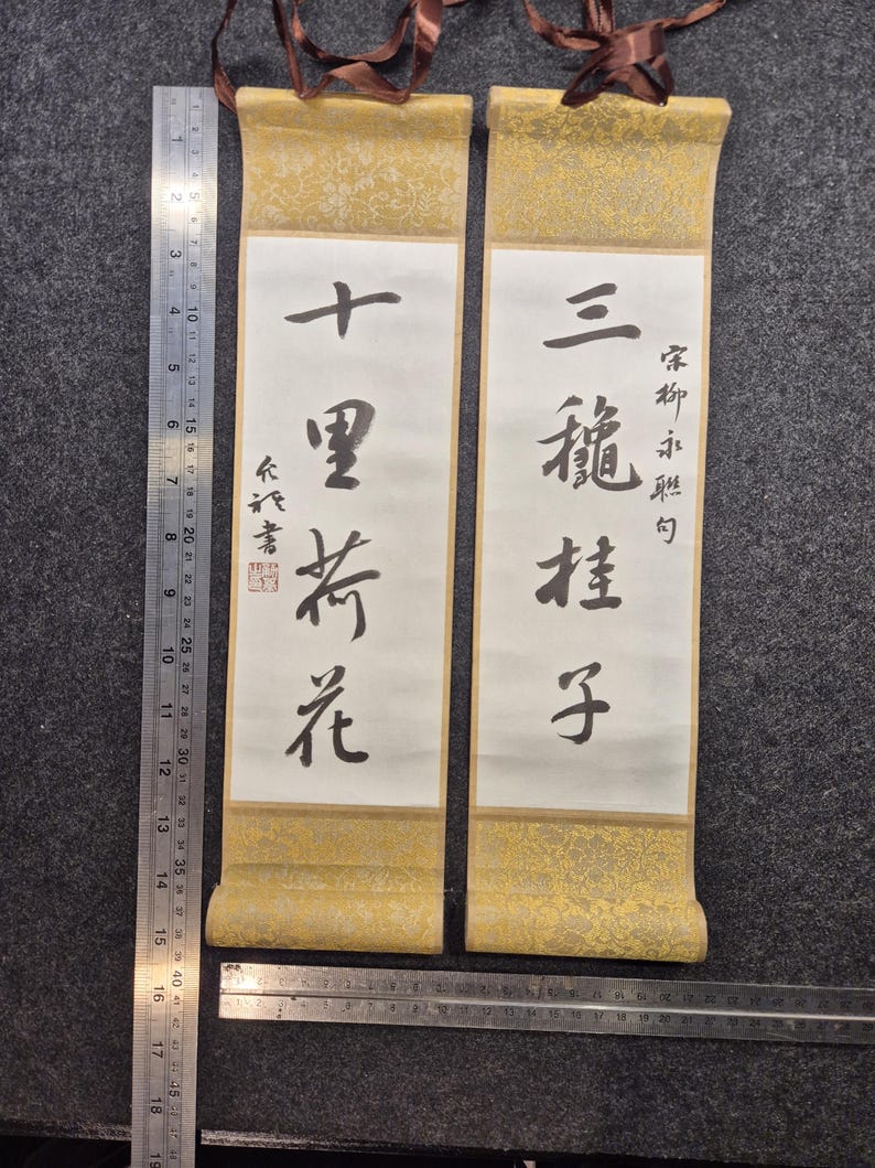 May include: Two traditional Asian scroll paintings. Each scroll features calligraphy on white paper, framed by gold patterned borders. The scrolls are suspended by brown ribbons. A ruler is placed next to the scrolls.