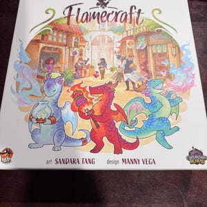 May include: A board game box cover with the title "Flamecraft" in a stylized font. The cover features a whimsical illustration of three dragons in a medieval town setting. The dragons are blue, red, and green, and they are holding various objects. The text "art SANDARA TANG design MANNY VEGA" is at the bottom of the box.