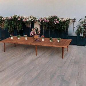 May include: A long, rectangular wooden table with a warm brown finish, set with small potted plants and a basket of flowers. The table is surrounded by greenery and flowers, creating a natural and inviting atmosphere. The table's legs are a darker brown.