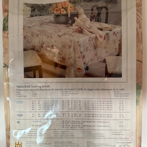 May include: A Bella Rosa tablecloth in a package. The tablecloth has a floral pattern with red and pink roses on a cream background. The packaging includes a tablecloth seating guide with measurements in inches and centimeters.