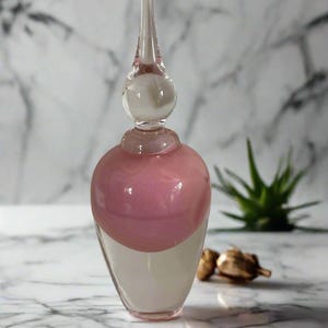 May include: A decorative glass perfume bottle with a long, pointed stopper. The bottle has a rounded base with a pink and white swirl design. The top of the bottle is clear glass. The bottle is on a marble surface.