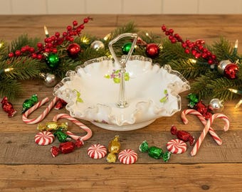 Puede incluir: Una fuente blanca de dos niveles con borde ondulado y asa plateada. El plato está decorado con un estampado floral y rodeado de bastones de caramelo, caramelos envueltos y adornos navideños. El fondo incluye una guirnalda de ramas de hoja perenne, bayas rojas y luces.
