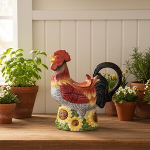 Peut inclure: Une théière en céramique en forme de coq avec une poignée noire et une base de tournesol. La théière est entourée d'herbes et de fleurs en pot, créant une esthétique rustique. La scène se déroule sur une table en bois près d'une fenêtre.