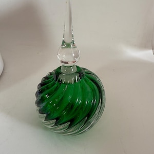 May include: A decorative glass perfume bottle with a clear, pointed stopper. The round body features a vibrant green and black swirl design. The bottle is made of clear glass.