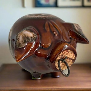 Pode incluir: Um cofrinho de cerâmica marrom com acabamento brilhante. O porco tem uma rolha de cortiça com um anel de metal. O banco tem uma ranhura na parte superior para inserir moedas e está sobre uma superfície de madeira.