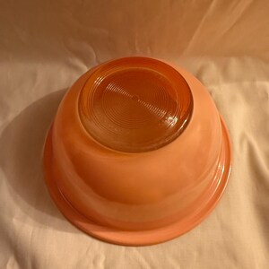 May include: A vintage, peach-colored glass mixing bowl, viewed from the bottom. The bowl has a rounded shape with a slightly flared rim. The base of the bowl features a circular, textured pattern. The bowl is set against a neutral background.