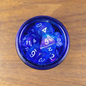 May include: A top-down view of a dark blue glass jar containing multiple blue and purple polyhedral dice. The dice, with white numerals, are for tabletop role-playing games. The jar rests on a wooden surface.