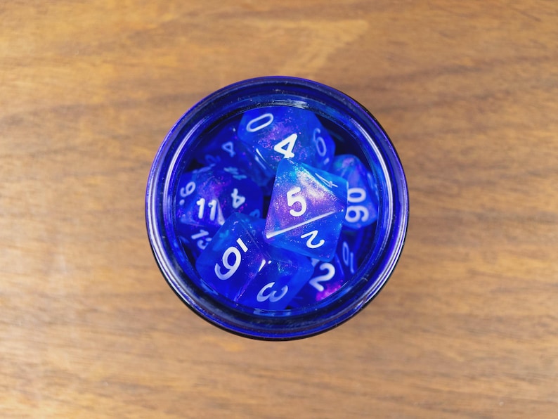 May include: A top-down view of a dark blue glass jar containing multiple blue and purple polyhedral dice. The dice, with white numerals, are for tabletop role-playing games. The jar rests on a wooden surface.