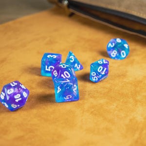 May include: A selection of blue and purple polyhedral dice with white numbering, scattered on a tan surface. A leather pouch is visible in the background. These dice are designed for tabletop role-playing games.