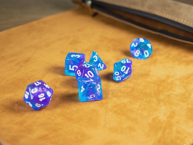 May include: A selection of blue and purple polyhedral dice with white numbering, scattered on a tan surface. A leather pouch is visible in the background. These dice are designed for tabletop role-playing games.