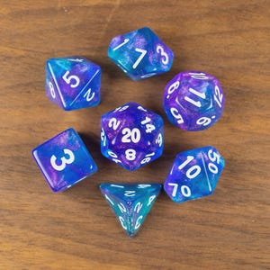 May include: A collection of seven multi-sided dice in blue and purple hues, each with white numerals. The dice are displayed on a wooden surface. These dice are designed for tabletop role-playing games.