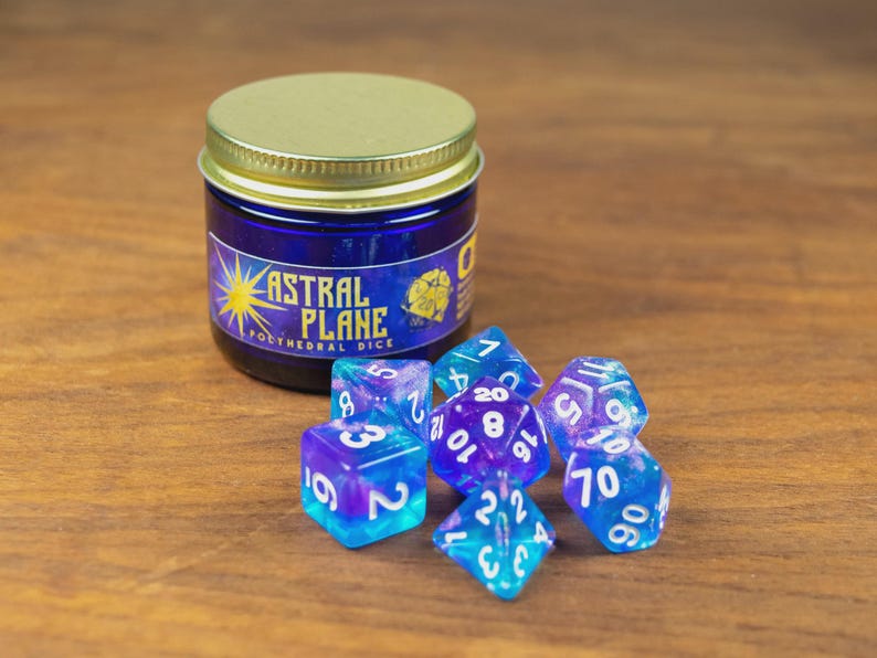 May include: A jar of blue dice with a gold lid and the text "ASTRAL PLANE POLYHEDRAL DICE". Several blue and purple polyhedral dice are scattered in front of the jar. The dice have white numbers.
