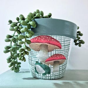 May include: A decorative plant pot with a green rim and a white and green checkered pattern. The pot features illustrations of red mushrooms and a green frog. A green plant cascades over the side of the pot.
