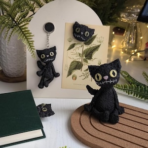 May include: A collection of handmade black crochet cat figures with stitched facial features. One cat is a keychain, another is a magnet, and a third is a small figurine. The cats have yellow eyes and a stitched smile. The background includes a botanical print and a green book.