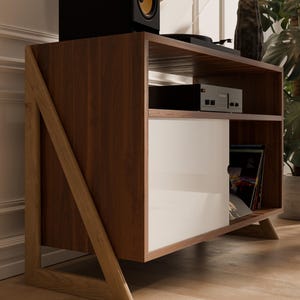 以下が含まれることがあります： 白いドアと茶色の木製のフレームが付いたミッドセンチュリーモダンなレコードプレーヤーのスタンド。スタンドにはレコード用の棚とターンテーブル用の上部棚があります。スタンドは明るい木製の床の上にあります。