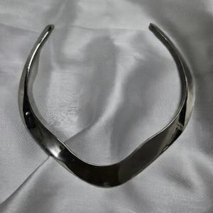 Könnte beinhalten: Eine silberfarbene Halskette mit glattem, geschwungenem Design. Die Halskette hat eine breite, flache Mitte, die sich zu den Enden hin verjüngt. Das Metall reflektiert das Licht und erzeugt Glanzlichter und Schatten. Die Halskette wird auf weißem, satinartigem Stoff präsentiert.