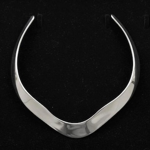 Peut inclure: Un collier ras du cou argenté poli avec un design lisse et incurvé. Le collier a un centre large et plat qui se rétrécit en extrémités plus fines. La pièce est présentée sur un fond noir, soulignant sa surface réfléchissante.