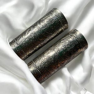 May include: A pair of silver arm cuffs with detailed floral and geometric embossed patterns. The cylindrical cuffs are displayed on a white, silky fabric. The intricate designs give a vintage or bohemian feel.