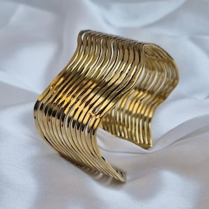 May include: A gold-colored cuff bracelet with a wavy design. The bracelet is made of multiple parallel, curved bars that create a textured, openwork appearance. The metal has a polished finish, reflecting light.