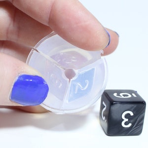 May include: A clear plastic dice rolling tray with three sections, each with a number printed on it. The numbers are 2, 3, and 5. A black dice with white numbers is sitting on a white surface.